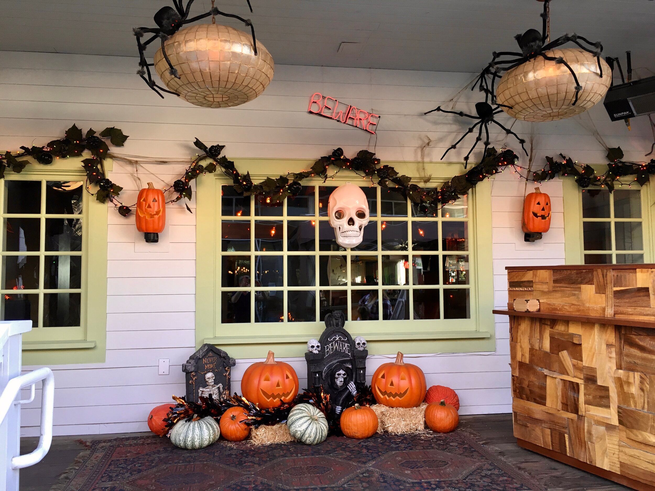 A porch decorated for Halloween with giant hanging spiders, jack-o'-lanterns and a lit BEWARE sign